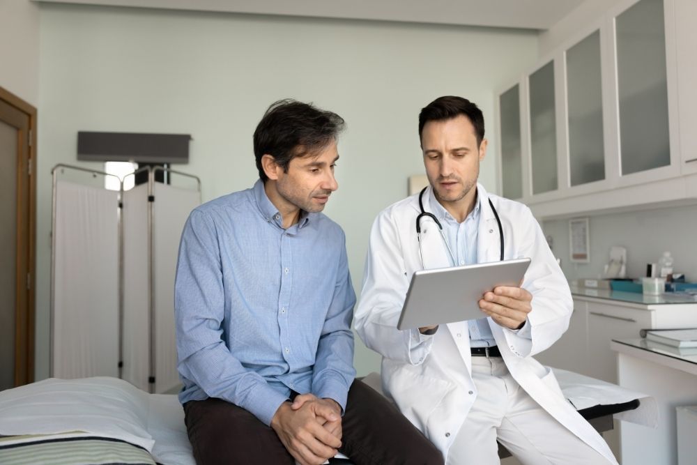 A doctor in a white coat shows a male patient his medical results on a digital tablet in an examination room.