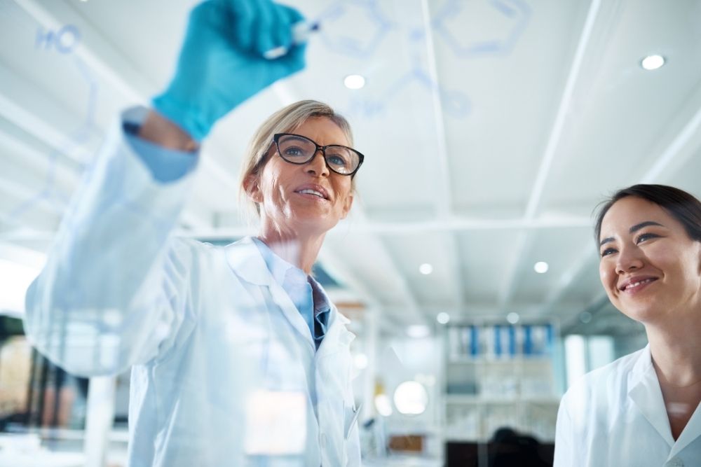 Two female scientists in lab coats, one writing chemical structures on a transparent glass board with a marker while the other watches with a smile.