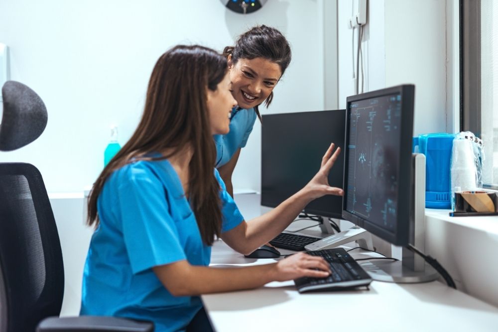 Two female nurses in blue scrubs smile while working together at a computer and analysing data on the screen in a modern clinic.