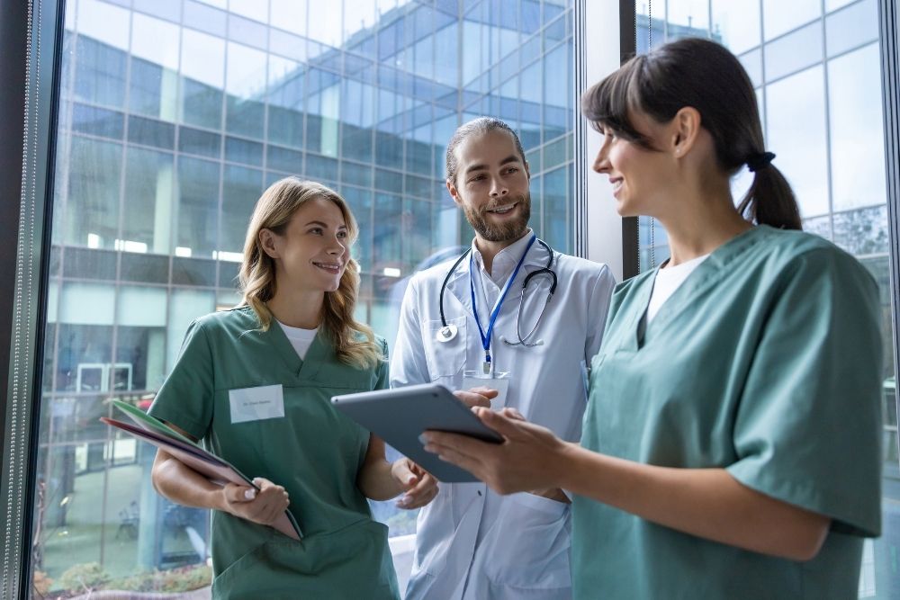 Medical-Team-Collaboration A doctor and two nurses smile as they discuss information on a digital tablet in a modern hospital hallway.