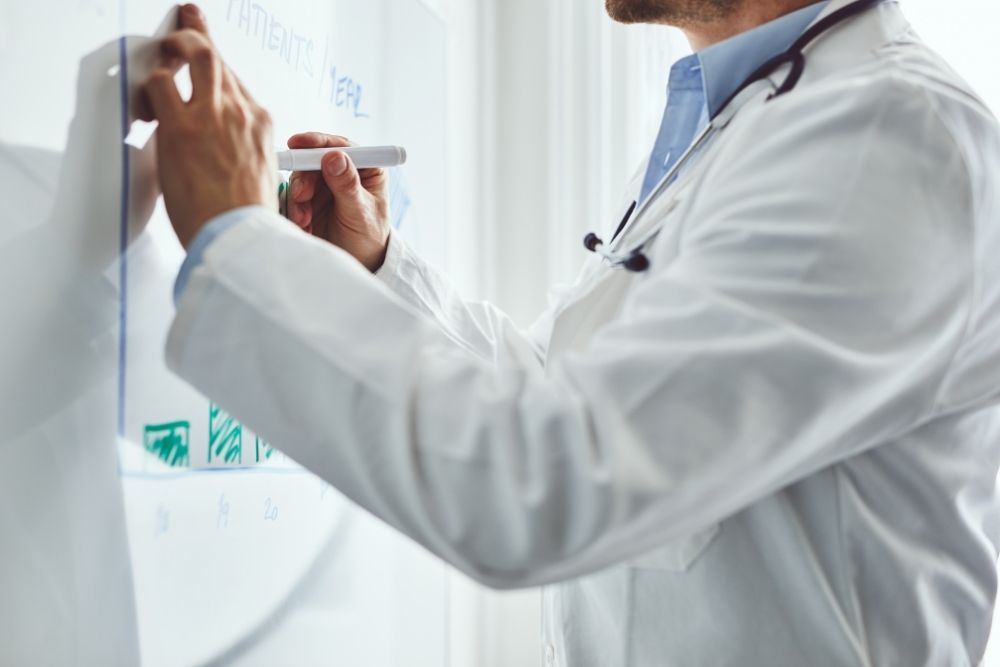 doctor-in-lab-coat-writing-on-a-whiteboard-during-a-presentation A doctor with a stethoscope wearing a white lab coat writes on a whiteboard with a marker.