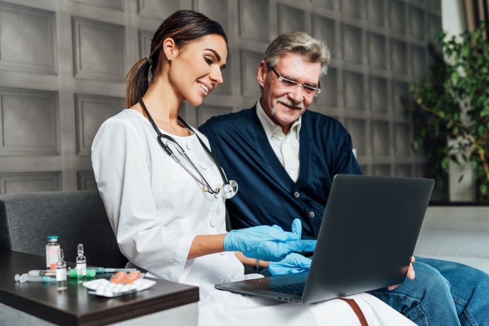 female-doctor-showing-elderly-male-patient-information-on-a-laptop A female doctor in a white coat shows an elderly male patient something on a laptop.