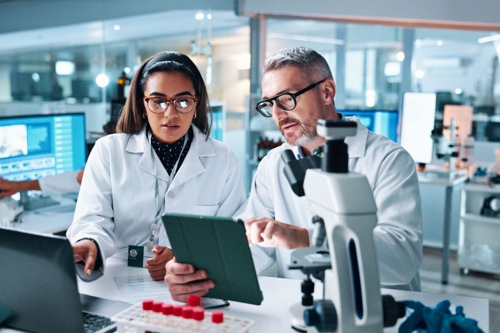 male-and-female-researchers-discussing-data-on-tablet-in-lab Two scientists in lab coats collaborating in a laboratory, looking at a tablet computer.